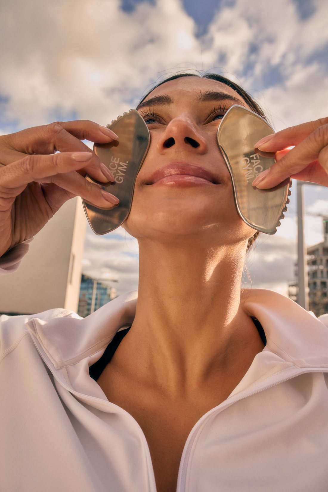 Maria holding two gua sha tools to her face against a cloudy sky